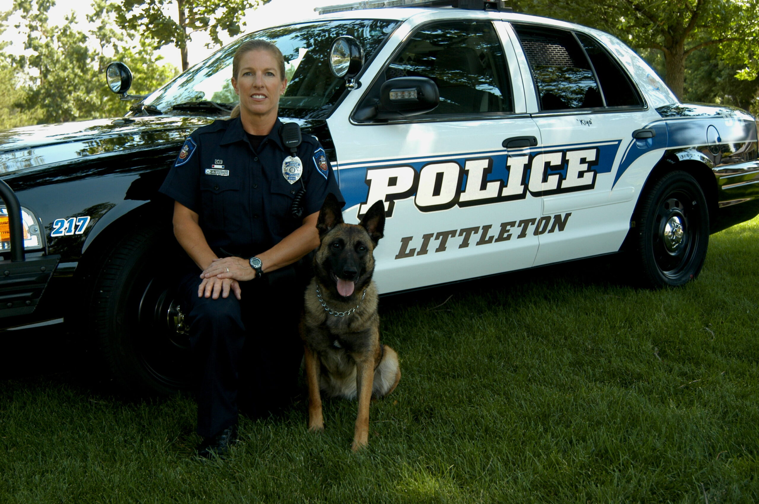 Police officer kneels beside a police dog in front of a patrol car.