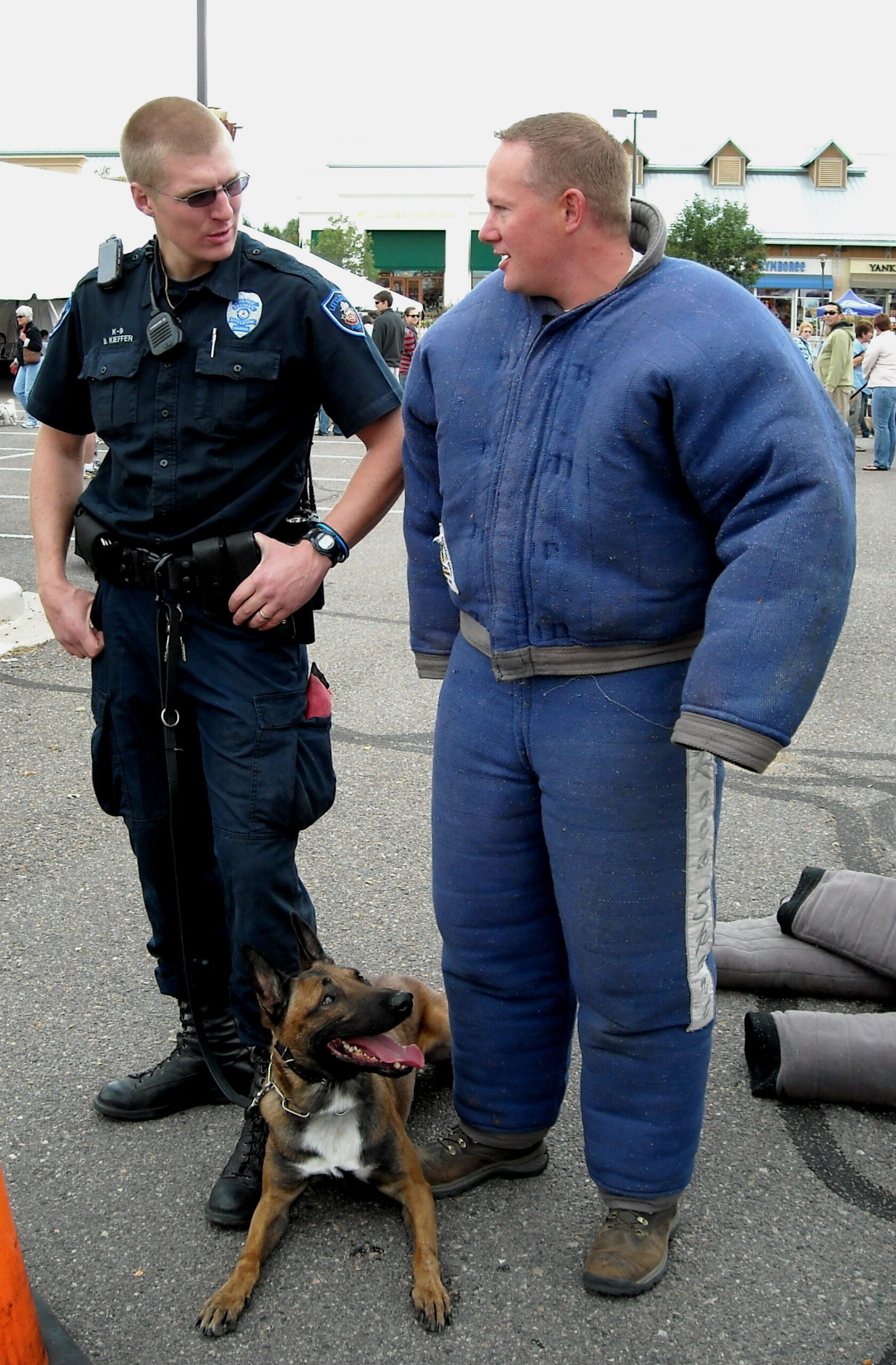 A police officer stands next to a person in a thick protective suit with a dog.