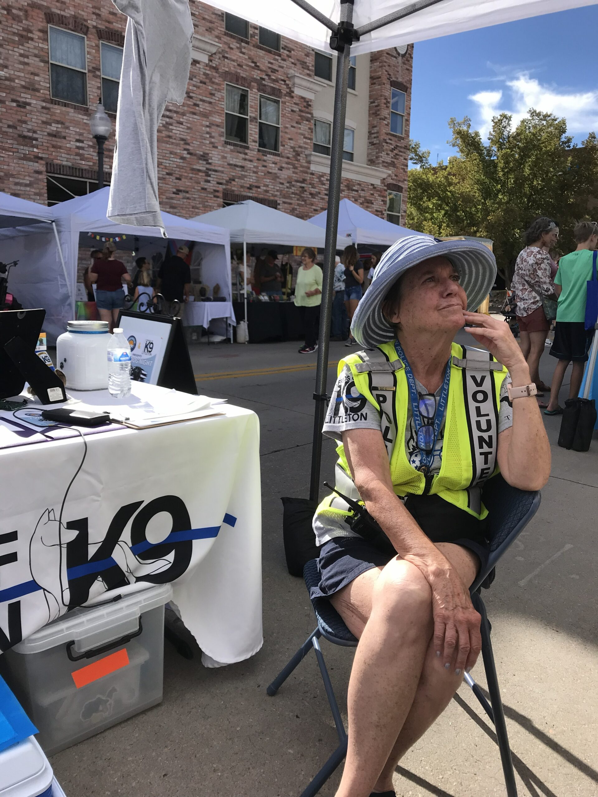 Woman in a yellow vest sitting by a K-9 unit table at an outdoor event.