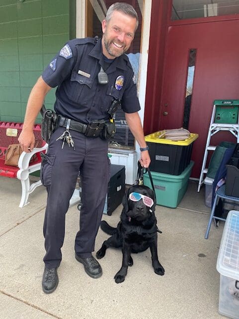 Police officer kneeling beside a black dog wearing sunglasses.
